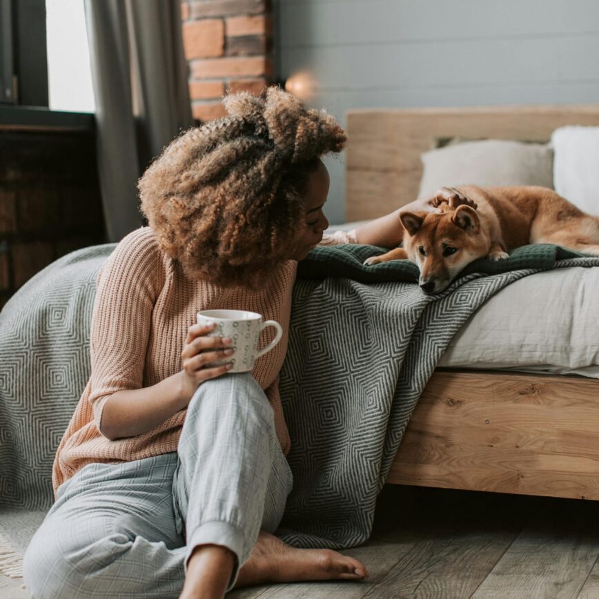 a woman sat on the floor against a bed whilst holding a mug and patting her dog lay behind her