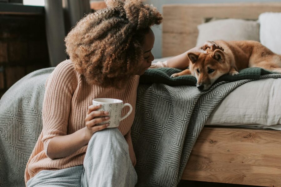 a woman sat on the floor against a bed whilst holding a mug and patting her dog lay behind her