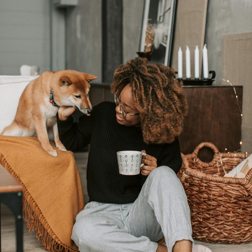 a woman sat on the floor holding a mug whilst patting her dog sat beside her.