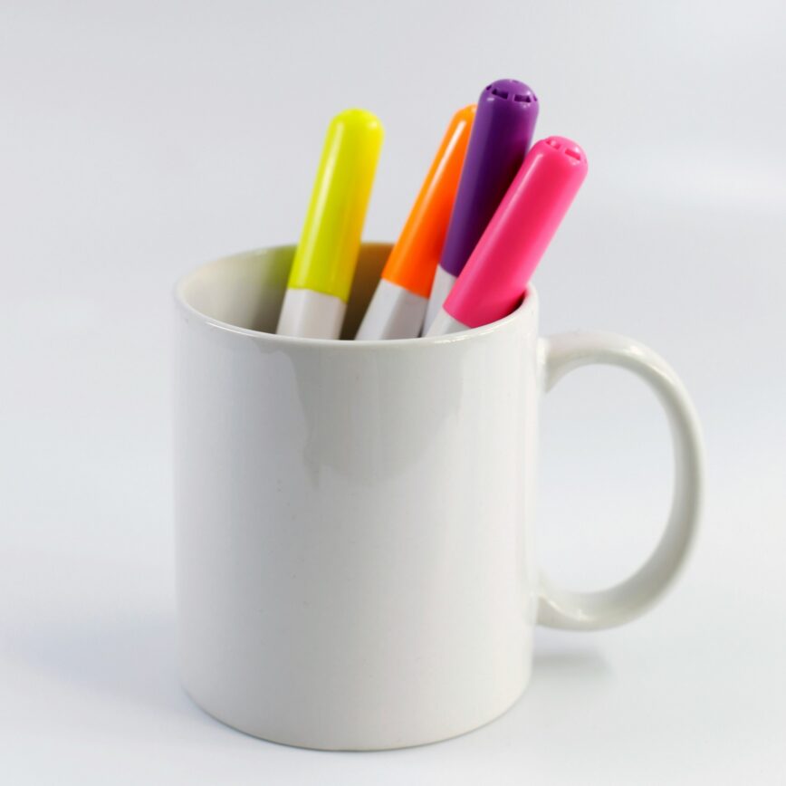 a white ceramic mug with various coloured pens being stored within it.