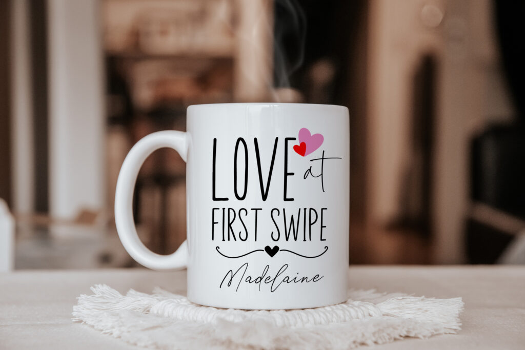 

a ceramic mug pictured on a white table, the design upon the mug shares the wording 'love at first swipe' with a personalised name and a floating heart watercolour illustration.